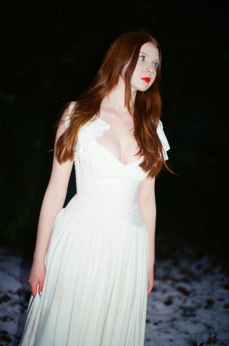 May include: A woman in a white dress with a fitted bodice and flowing skirt stands outdoors. She has long auburn hair and red lipstick. The dress has a sweetheart neckline and ruffled sleeves. The background is dark with a hint of snow.