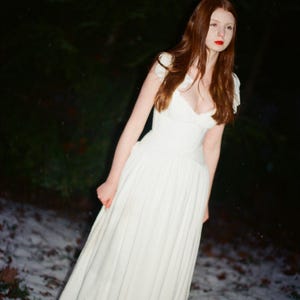 May include: A woman in a white dress with a low neckline and cap sleeves stands outdoors. The long dress has a pleated skirt. The woman has long auburn hair and red lipstick. The background is a dark forest with snow on the ground.