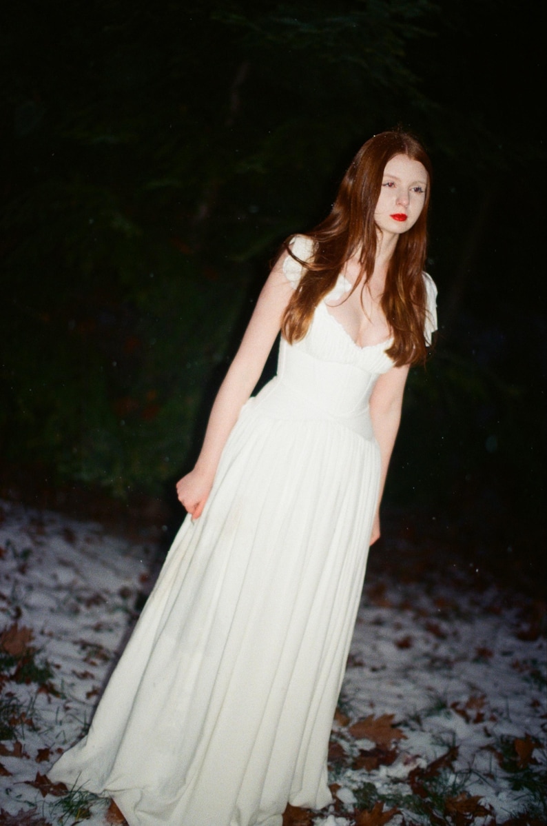 May include: A woman in a white dress with a low neckline and cap sleeves stands outdoors. The long dress has a pleated skirt. The woman has long auburn hair and red lipstick. The background is a dark forest with snow on the ground.