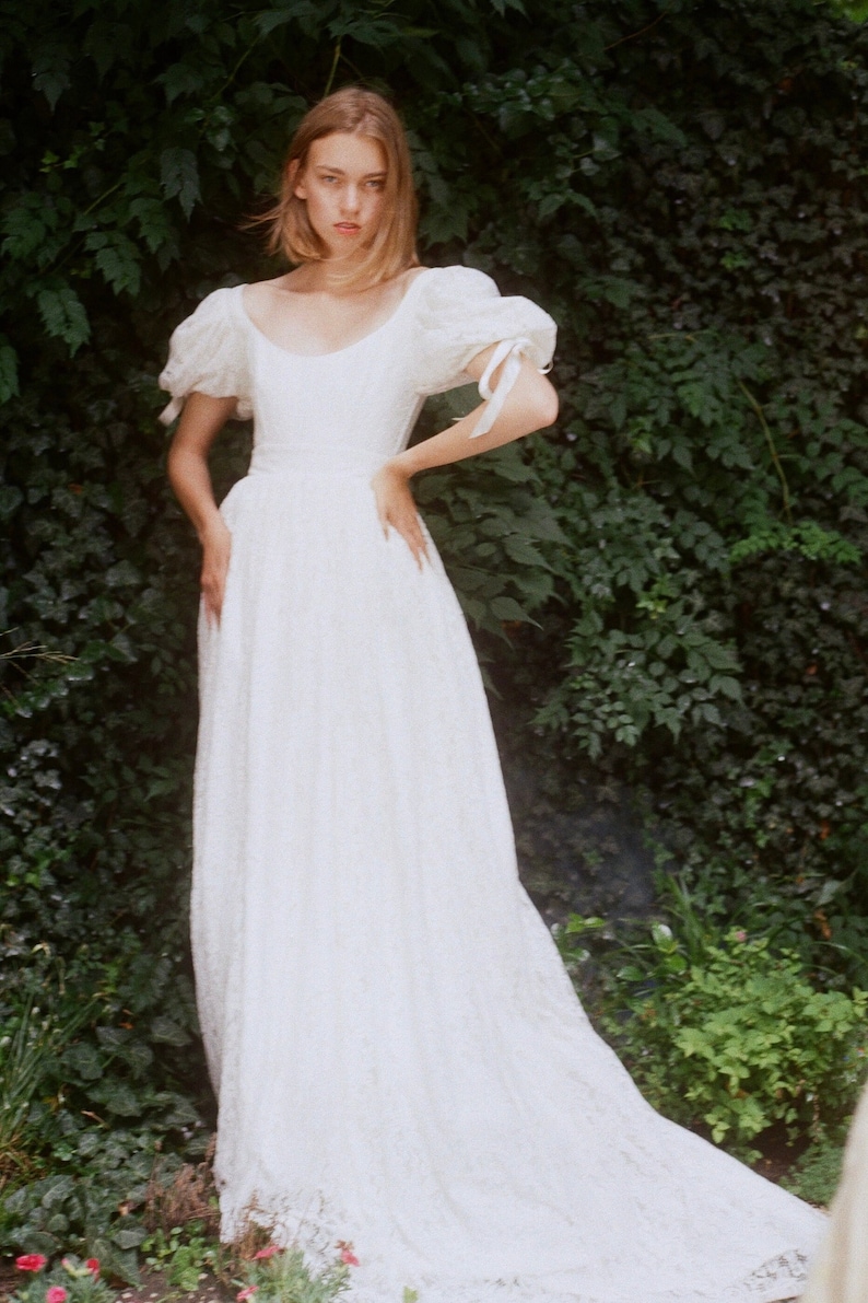 May include: A white lace dress with a scoop neckline and puffed sleeves. The dress has a fitted bodice and a flowing skirt. The dress is worn by a person standing in front of a green leafy background.