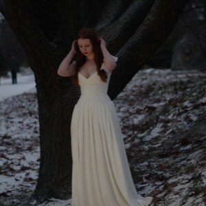 May include: A long, white dress with a fitted bodice and flowing skirt. The dress has a low neckline and short sleeves. The person is standing in a snowy, wooded area with a large tree in the background.