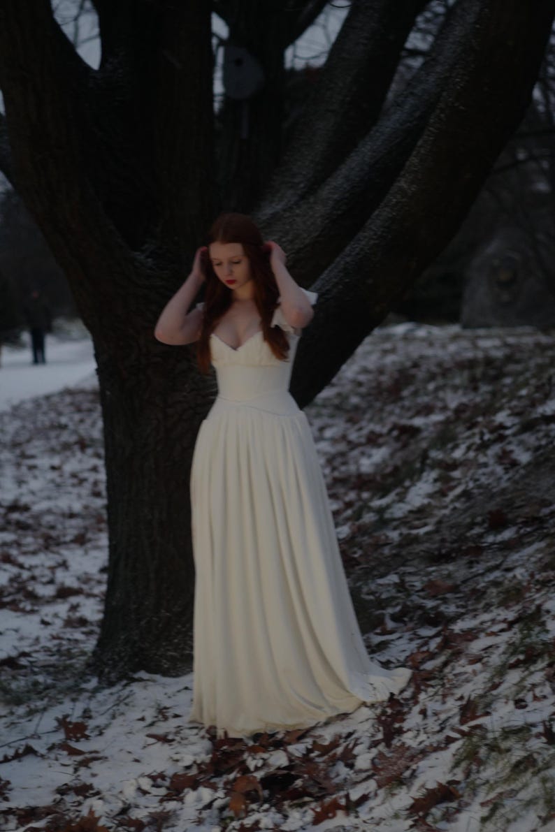 May include: A long, white dress with a fitted bodice and flowing skirt. The dress has a low neckline and short sleeves. The person is standing in a snowy, wooded area with a large tree in the background.