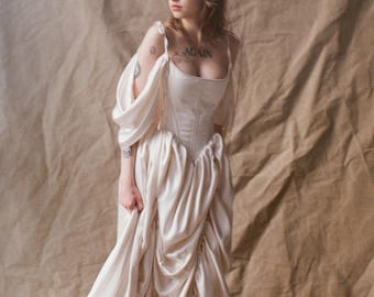 May include: A floor-length cream-colored gown featuring a structured corset-style bodice with a square neckline and thin shoulder straps. The skirt is voluminous with gathered fabric detailing, and the sleeves are loose, draped, and off-the-shoulder.