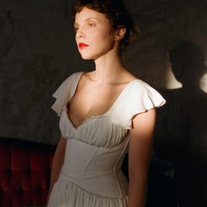 May include: A white dress with a fitted bodice and flowing skirt. The dress features a sweetheart neckline and ruffled sleeves. The woman in the image has red lipstick and is lit by sunlight, creating a dramatic effect. The dress is likely for a special occasion.