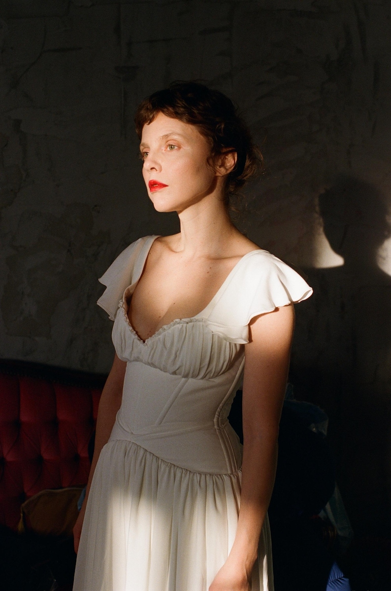 May include: A white dress with a fitted bodice and flowing skirt. The dress features a sweetheart neckline and ruffled sleeves. The woman in the image has red lipstick and is lit by sunlight, creating a dramatic effect. The dress is likely for a special occasion.