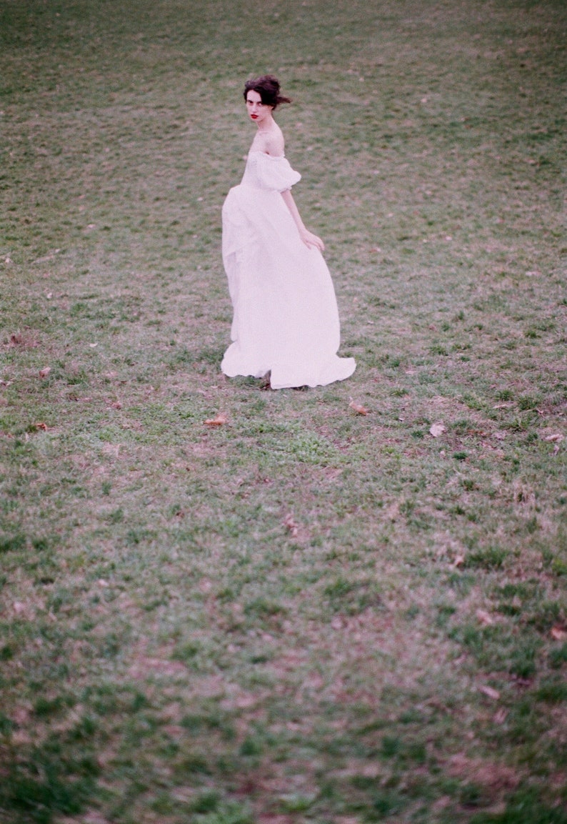 Puede incluir: Una mujer con un vestido blanco sin tirantes se encuentra en un campo de hierba. Est&aacute; mirando a la c&aacute;mara y tiene el pelo recogido hacia atr&aacute;s.