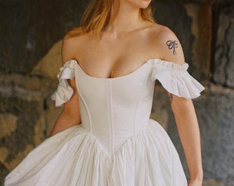 Puede incluir: Un vestido blanco de hombros descubiertos con un corpiño estructurado estilo corsé con costuras verticales y escote corazón. La prenda incluye mangas cortas con volantes y una falda voluminosa fruncida que cae desde la cintura.