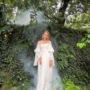 Puede incluir: Una mujer con un vestido blanco de hombros descubiertos con mangas abullonadas se encuentra en un jard&iacute;n con humo a su alrededor. El vestido tiene un corpi&ntilde;o ajustado y una falda fluida.