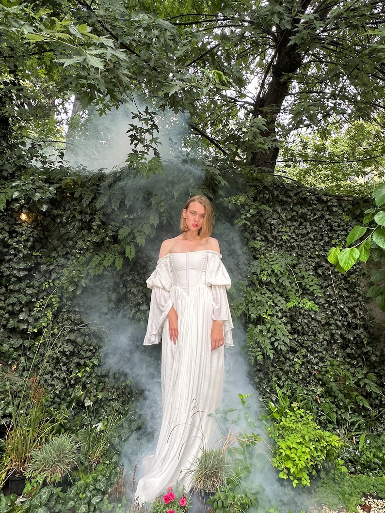Puede incluir: Una mujer con un vestido blanco de hombros descubiertos con mangas abullonadas se encuentra en un jard&iacute;n con humo a su alrededor. El vestido tiene un corpi&ntilde;o ajustado y una falda fluida.