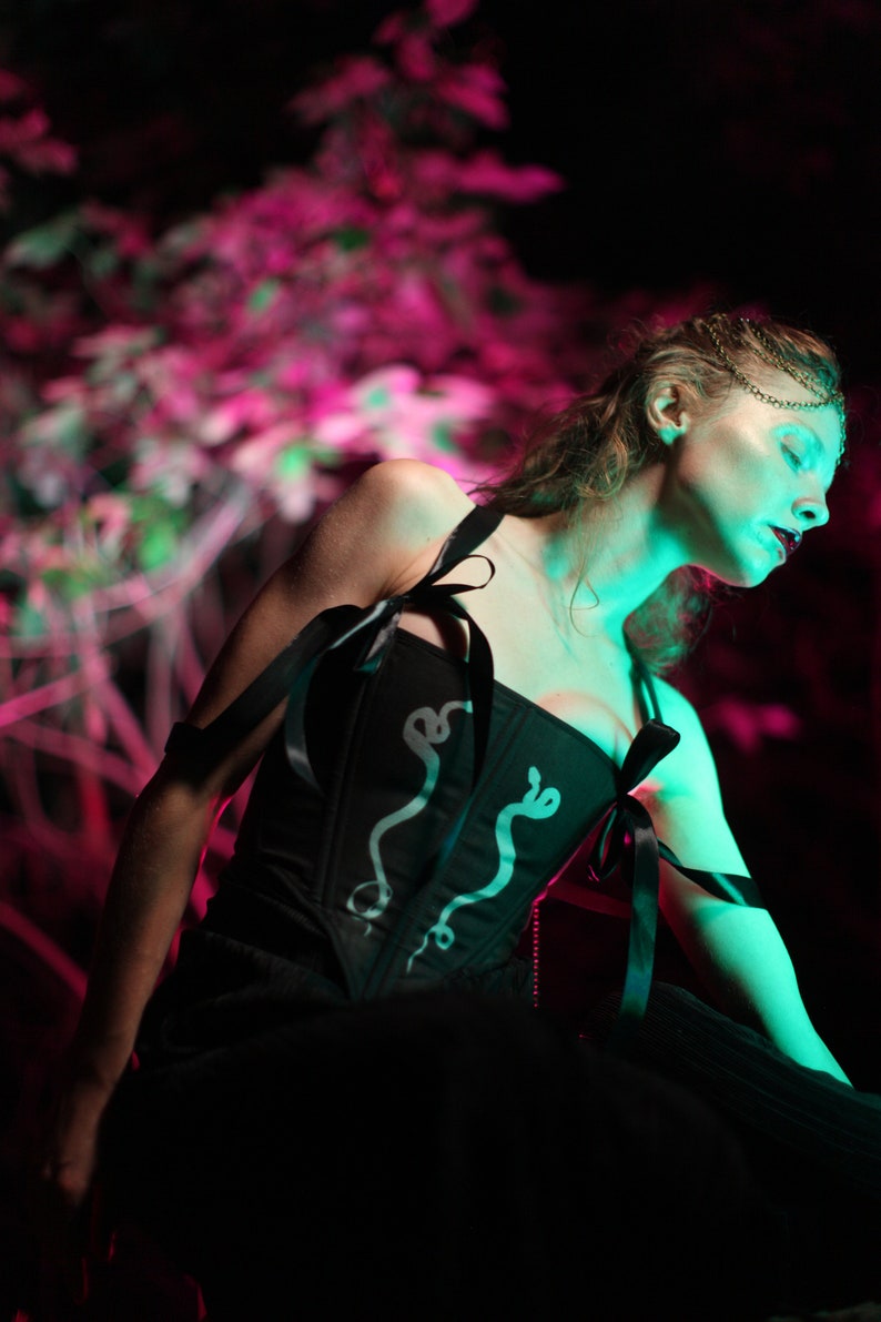 May include: A woman wearing a black corset with a snake design and a silver chain. The corset has black ribbon ties. The woman is standing in front of a pink and green blurred background.