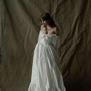 May include: A white off-the-shoulder dress featuring a fitted bodice, long puff sleeves with ruffled cuffs, and a full-length skirt with a ruffled hem. The garment has a sweetheart neckline and a central tie detail at the chest area.