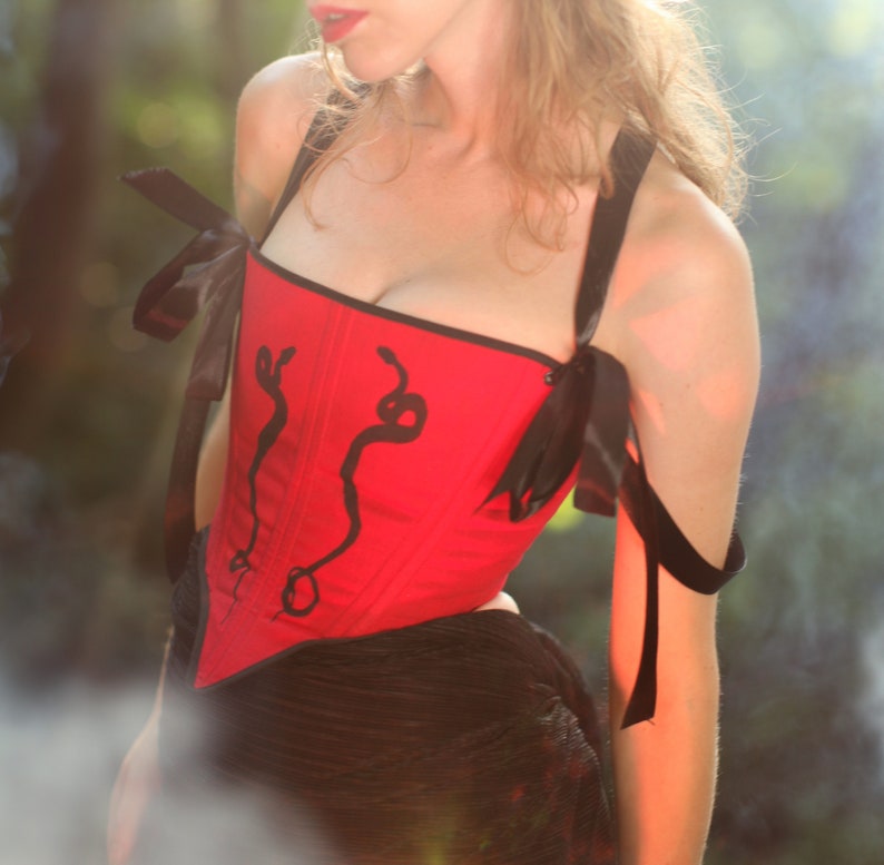 May include: A red corset with black ribbon straps and a snake design. The corset is worn by a person with long blonde hair.