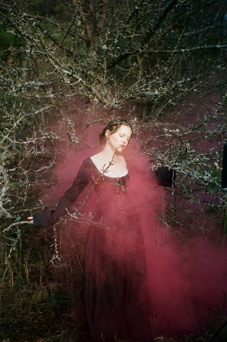 May include: A long-sleeved black dress with a scoop neckline and a corset-style bodice featuring front lacing. The garment is surrounded by a dense cloud of deep pink smoke, set against a backdrop of bare, lichen-covered tree branches.