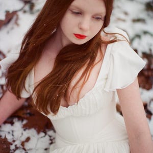 May include: A woman with long, auburn hair and red lipstick wears a white dress with ruffled sleeves. The dress has a fitted bodice and a flowing skirt. The background shows snow and fallen leaves, creating a winter scene.