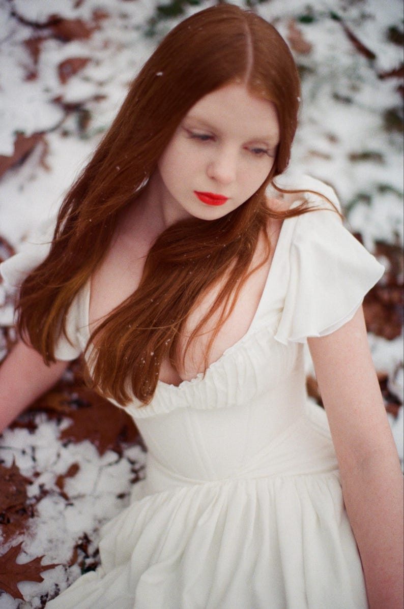 May include: A woman with long, auburn hair and red lipstick wears a white dress with ruffled sleeves. The dress has a fitted bodice and a flowing skirt. The background shows snow and fallen leaves, creating a winter scene.
