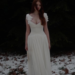 May include: A white dress with a low neckline and ruffled sleeves. The dress has a fitted bodice and a flowing skirt. The person in the image has long red hair and is standing in a snowy forest setting.