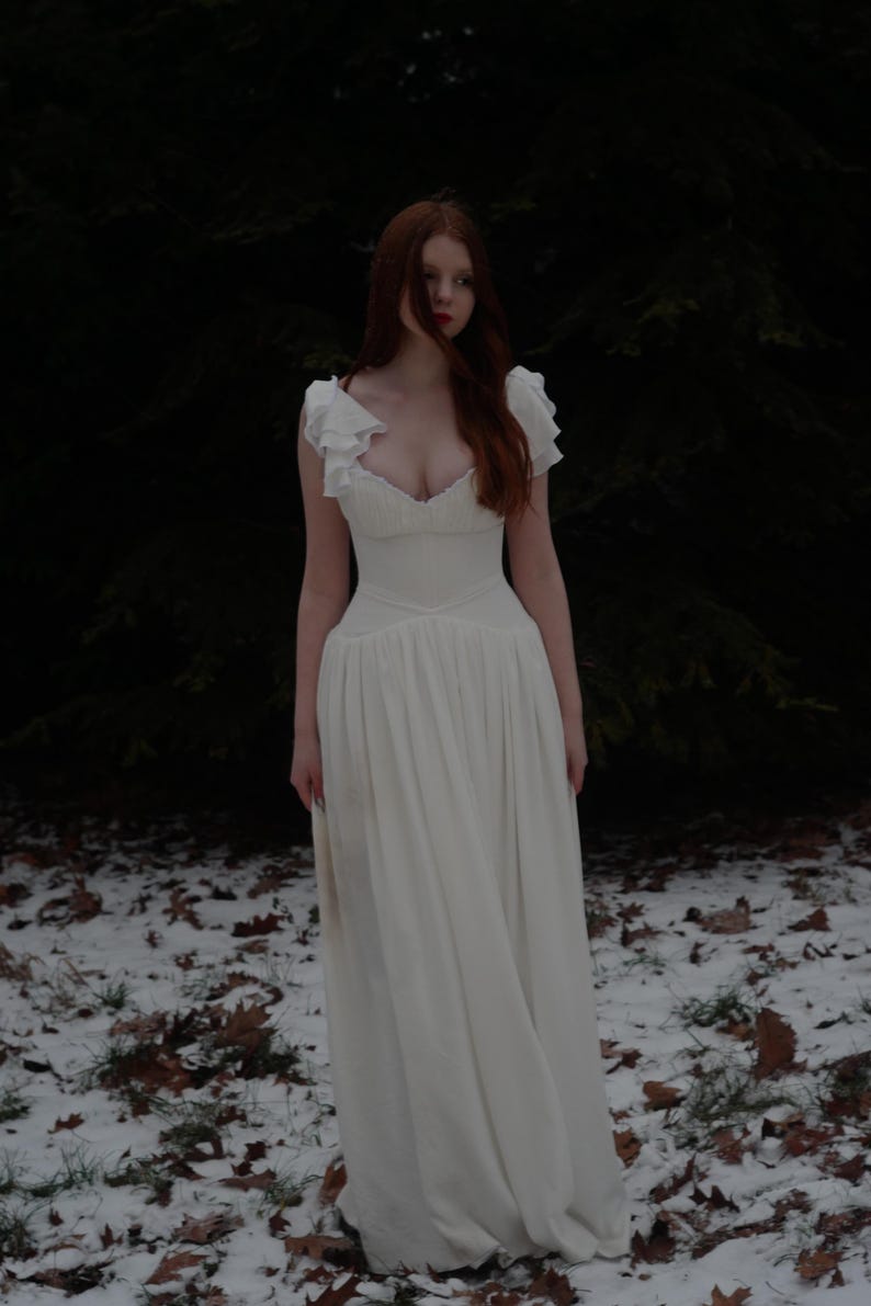 May include: A white dress with a low neckline and ruffled sleeves. The dress has a fitted bodice and a flowing skirt. The person in the image has long red hair and is standing in a snowy forest setting.