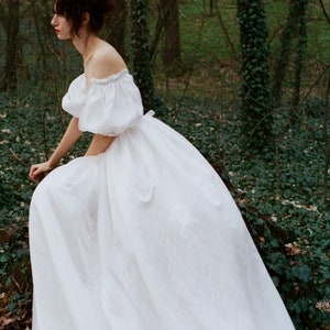 Puede incluir: Una mujer con un vestido blanco de hombros descubiertos y mangas abullonadas posa en un entorno de bosque. El vestido es fluido y ondea con la brisa.