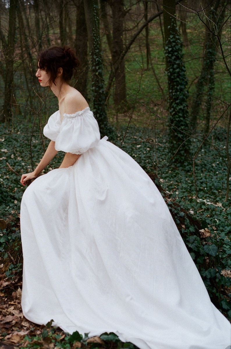 Puede incluir: Una mujer con un vestido blanco de hombros descubiertos y mangas abullonadas posa en un entorno de bosque. El vestido es fluido y ondea con la brisa.