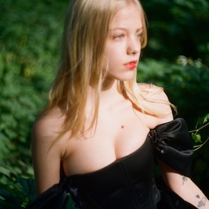 May include: A woman with long blonde hair wearing a black off-the-shoulder corset top with a ruffled skirt. She is standing in a forest setting.