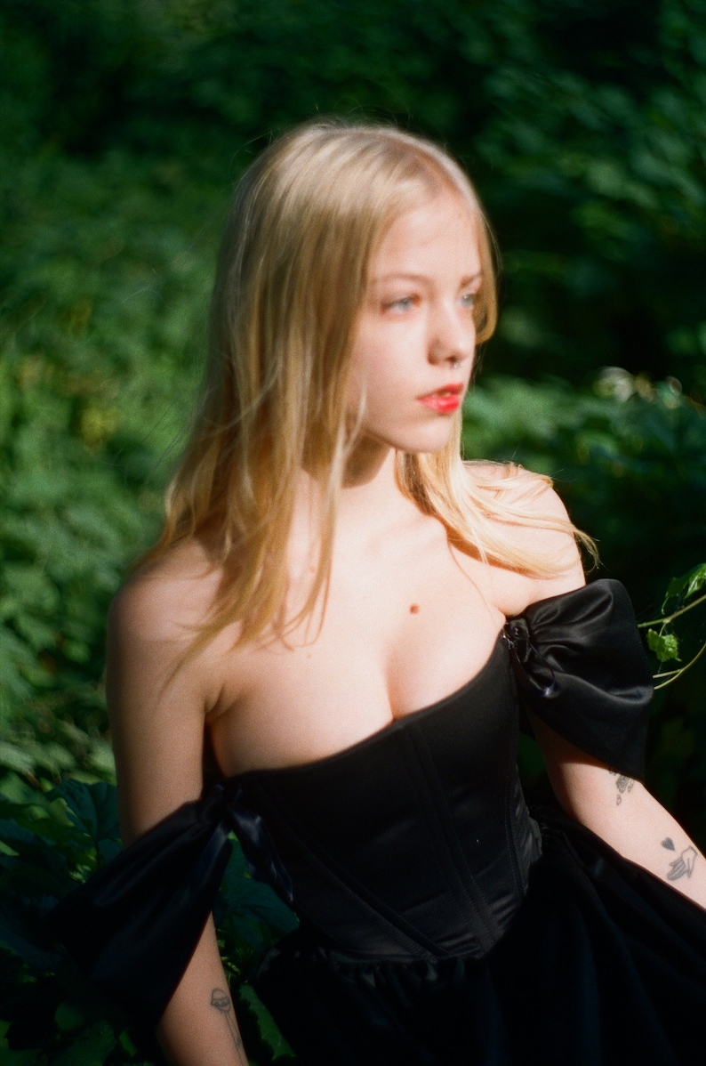 May include: A woman with long blonde hair wearing a black off-the-shoulder corset top with a ruffled skirt. She is standing in a forest setting.