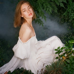 Puede incluir: Una mujer con un vestido blanco de hombros descubiertos se sienta en un jard&iacute;n con vegetaci&oacute;n exuberante y flores rosas. Tiene el pelo largo y casta&ntilde;o y mira a la c&aacute;mara.