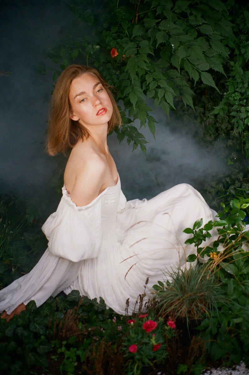 Puede incluir: Una mujer con un vestido blanco de hombros descubiertos se sienta en un jard&iacute;n con vegetaci&oacute;n exuberante y flores rosas. Tiene el pelo largo y casta&ntilde;o y mira a la c&aacute;mara.