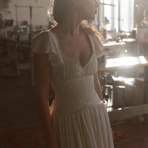 May include: A white dress with a fitted bodice and flowing skirt. The dress features a ruffled neckline and cap sleeves. The image is taken in a sunlit room, highlighting the dress's details and silhouette. The dress appears to be made of a soft, lightweight fabric.