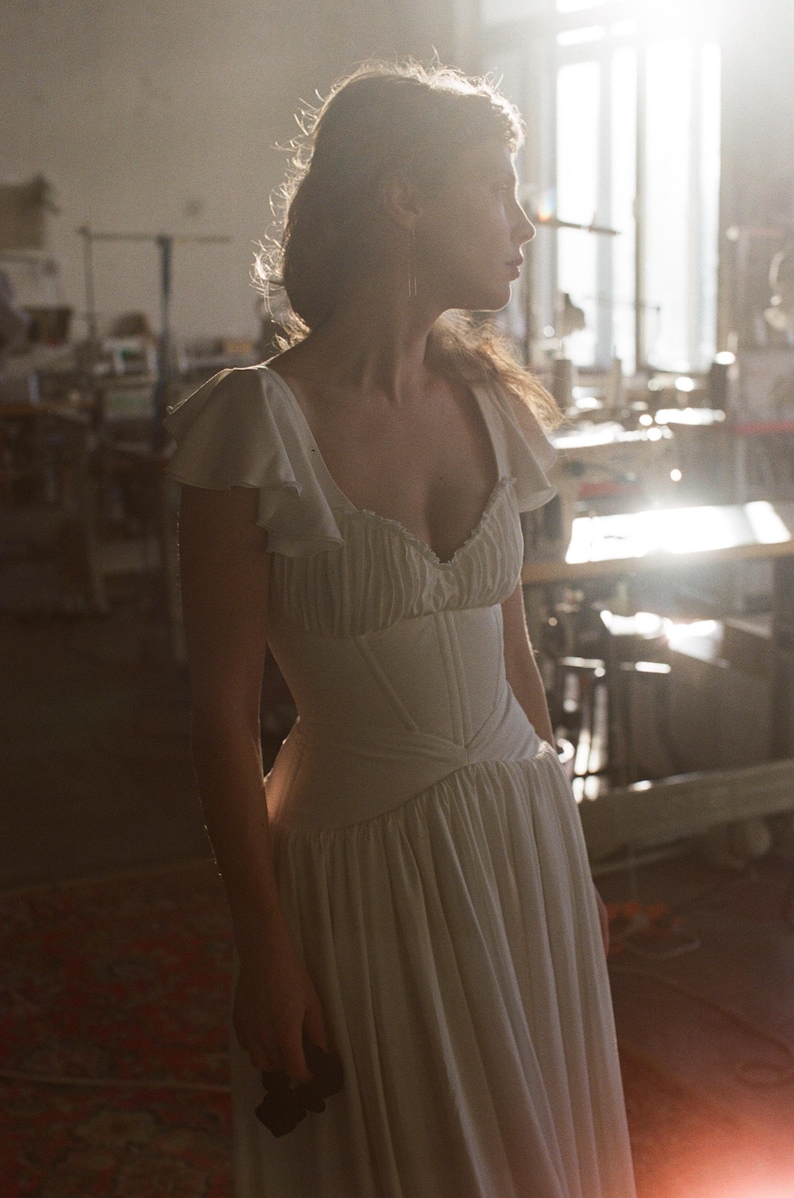 May include: A white dress with a fitted bodice and flowing skirt. The dress features a ruffled neckline and cap sleeves. The image is taken in a sunlit room, highlighting the dress's details and silhouette. The dress appears to be made of a soft, lightweight fabric.