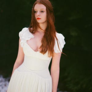 May include: A woman in a white dress with a low neckline and short, ruffled sleeves. The dress has a fitted bodice and a flowing skirt. The woman has long red hair and is standing in front of a dark green background with some snow on the ground.