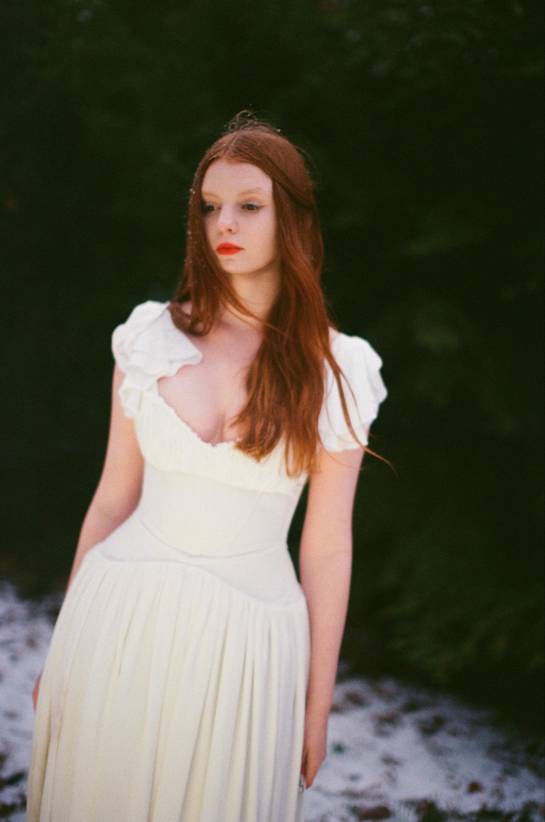 May include: A woman in a white dress with a low neckline and short, ruffled sleeves. The dress has a fitted bodice and a flowing skirt. The woman has long red hair and is standing in front of a dark green background with some snow on the ground.