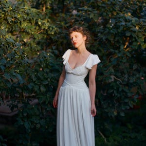 May include: A white dress with a fitted bodice and flowing skirt. The dress features a sweetheart neckline and cap sleeves. The image is set in a garden with lush green foliage, creating a soft, romantic aesthetic.