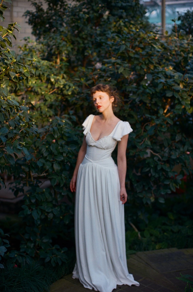May include: A white dress with a fitted bodice and flowing skirt. The dress features a sweetheart neckline and cap sleeves. The image is set in a garden with lush green foliage, creating a soft, romantic aesthetic.