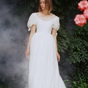 May include: A woman wearing a white lace dress with puffed sleeves. The dress has a fitted bodice and a flowing skirt. The woman is standing in a garden with green foliage and pink flowers.
