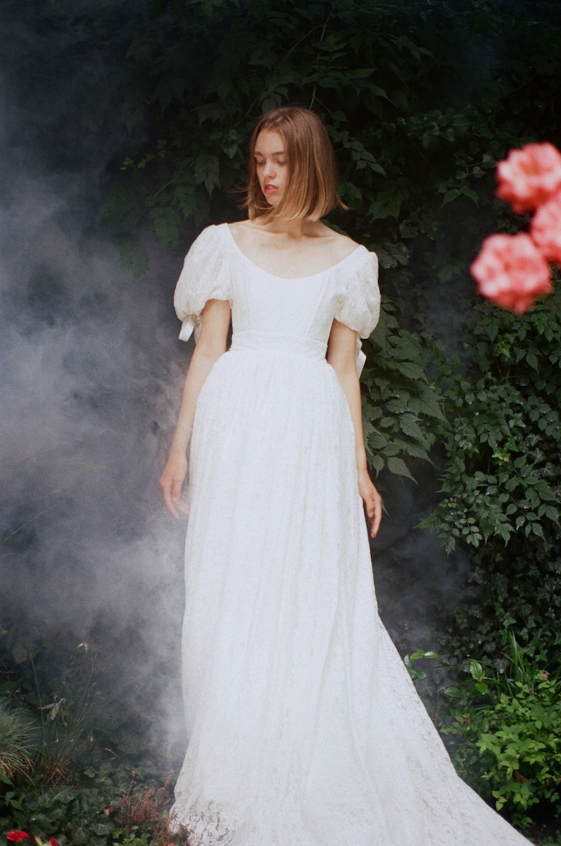 May include: A woman wearing a white lace dress with puffed sleeves. The dress has a fitted bodice and a flowing skirt. The woman is standing in a garden with green foliage and pink flowers.