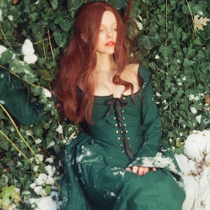 May include: A long-sleeved, floor-length dark green dress featuring a square neckline and a front bodice corset-style closure with a black ribbon lace-up detail and metal eyelets. The garment has a fitted bodice and a full, flowing skirt.