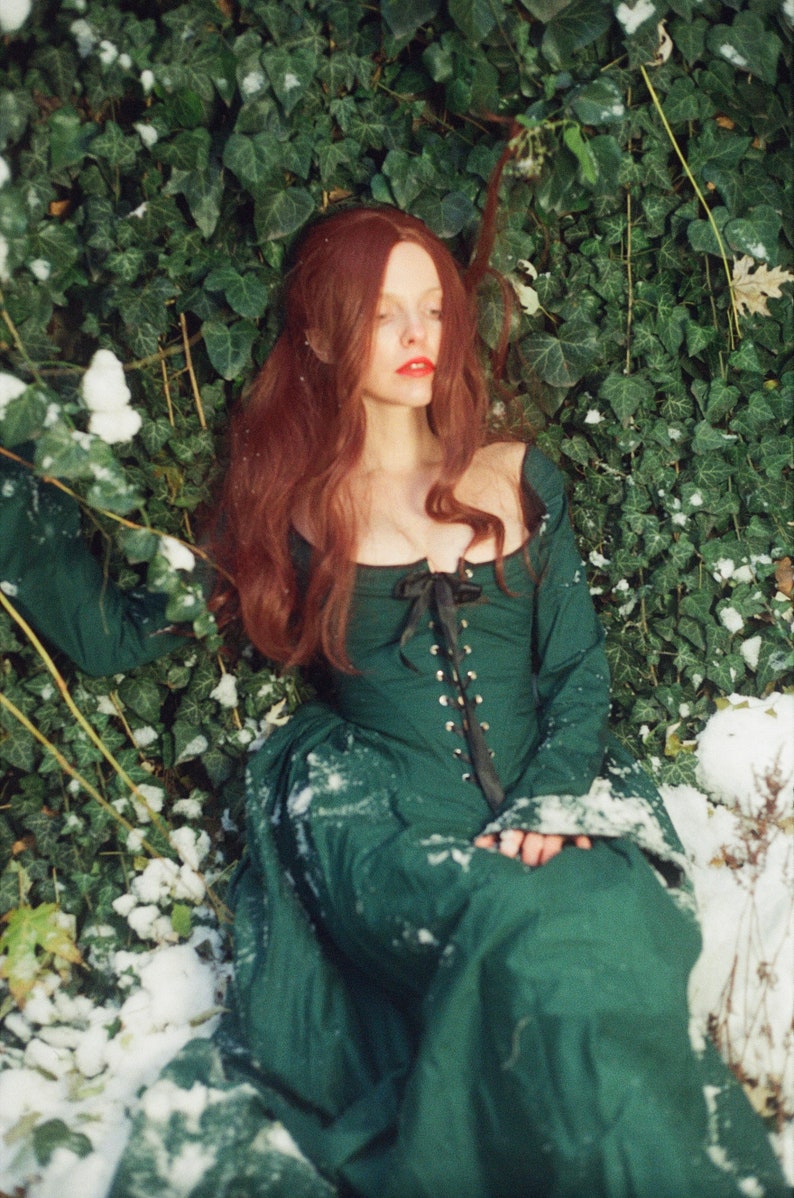 May include: A long-sleeved, floor-length dark green dress featuring a square neckline and a front bodice corset-style closure with a black ribbon lace-up detail and metal eyelets. The garment has a fitted bodice and a full, flowing skirt.