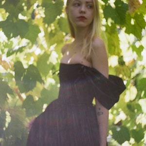 May include: A young woman with long blonde hair stands in a forest, wearing a black off-the-shoulder dress with a ruffled sleeve. The dress has a fitted bodice and a flowing skirt. The woman is looking off to the side, and the background is blurred.