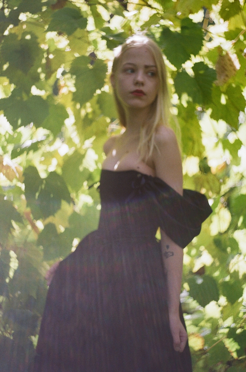 May include: A young woman with long blonde hair stands in a forest, wearing a black off-the-shoulder dress with a ruffled sleeve. The dress has a fitted bodice and a flowing skirt. The woman is looking off to the side, and the background is blurred.