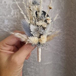 Könnte beinhalten: Ein kleiner Strauß getrockneter Blumen in Weiß- und Grautönen, gebunden mit einem weißen Band. Der Strauß enthält getrocknete Gräser, kleine weiße Blüten und graue Blüten.