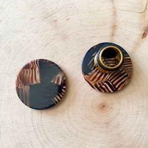 May include: Two-piece set of a bottle stopper and a coaster. The stopper has a gold-colored ring and a dark brown and tan patterned design. The coaster has a matching design. Both items are round and sit on a light wood surface.