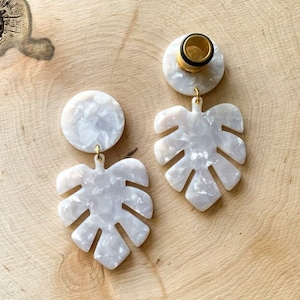 Könnte beinhalten: Ein Paar weiße, marmorierte, blattartige Ohrringe mit goldenen Akzenten. Die Ohrringe hängen an einer runden, weißen, marmorierten Scheibe.
