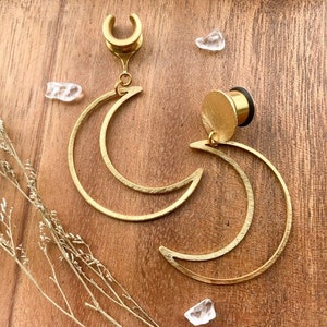 May include: A pair of gold crescent moon shaped earrings. The earrings are made of metal and have a smooth finish. The earrings are designed to be worn in stretched earlobes.