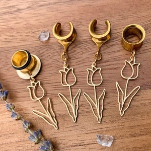 Pode incluir: Túneis de orelha dourados com um pingente de tulipa. Os túneis são feitos de metal e têm um acabamento polido. O pingente de tulipa é um contorno delicado da flor, com caule e folhas. Os túneis são exibidos em uma superfície de madeira.