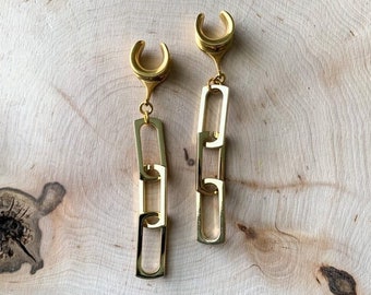 May include: A pair of gold-coloured ear gauges with a chain link design. The gauges have a curved top and a series of rectangular links that hang down. The jewellery is displayed on a light-coloured wooden surface.