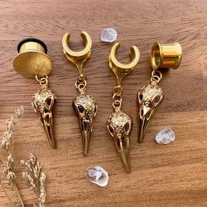 Puede incluir: Cuatro pendientes de calavera de color dorado con un diseño colgante. Los pendientes están hechos de metal y tienen un diseño de calavera detallado.