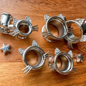 May include: Five silver cat-shaped ear gauges with rhinestones. The gauges are different sizes and are arranged on a wooden surface.