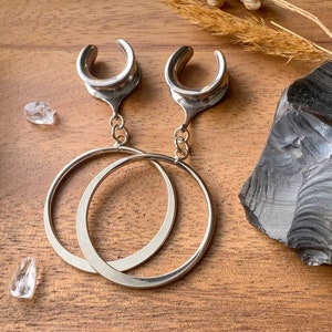 May include: A pair of silver hoop earrings with a dangling silver teardrop-shaped charm. The earrings are on a wooden surface.