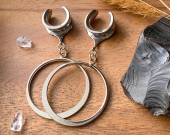 May include: A pair of silver hoop earrings with a dangling silver teardrop-shaped charm. The earrings are on a wooden surface.