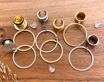 May include: Five pairs of gold and silver hoop earrings with different styles of ear gauges. The earrings are on a wooden surface.